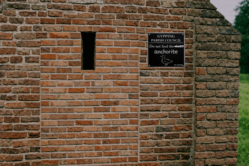 A brick-built cell attached to a church, with a bricked-up door; a sign is attached to the wall requesting that people do not feed the anchorite